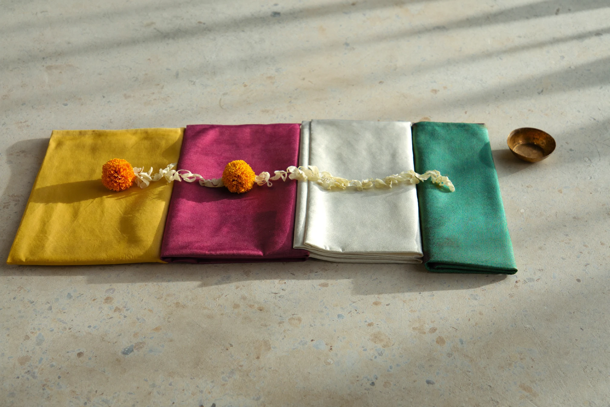 Four colourful folded silk textiles arranged with marigold petals and a brass diya.